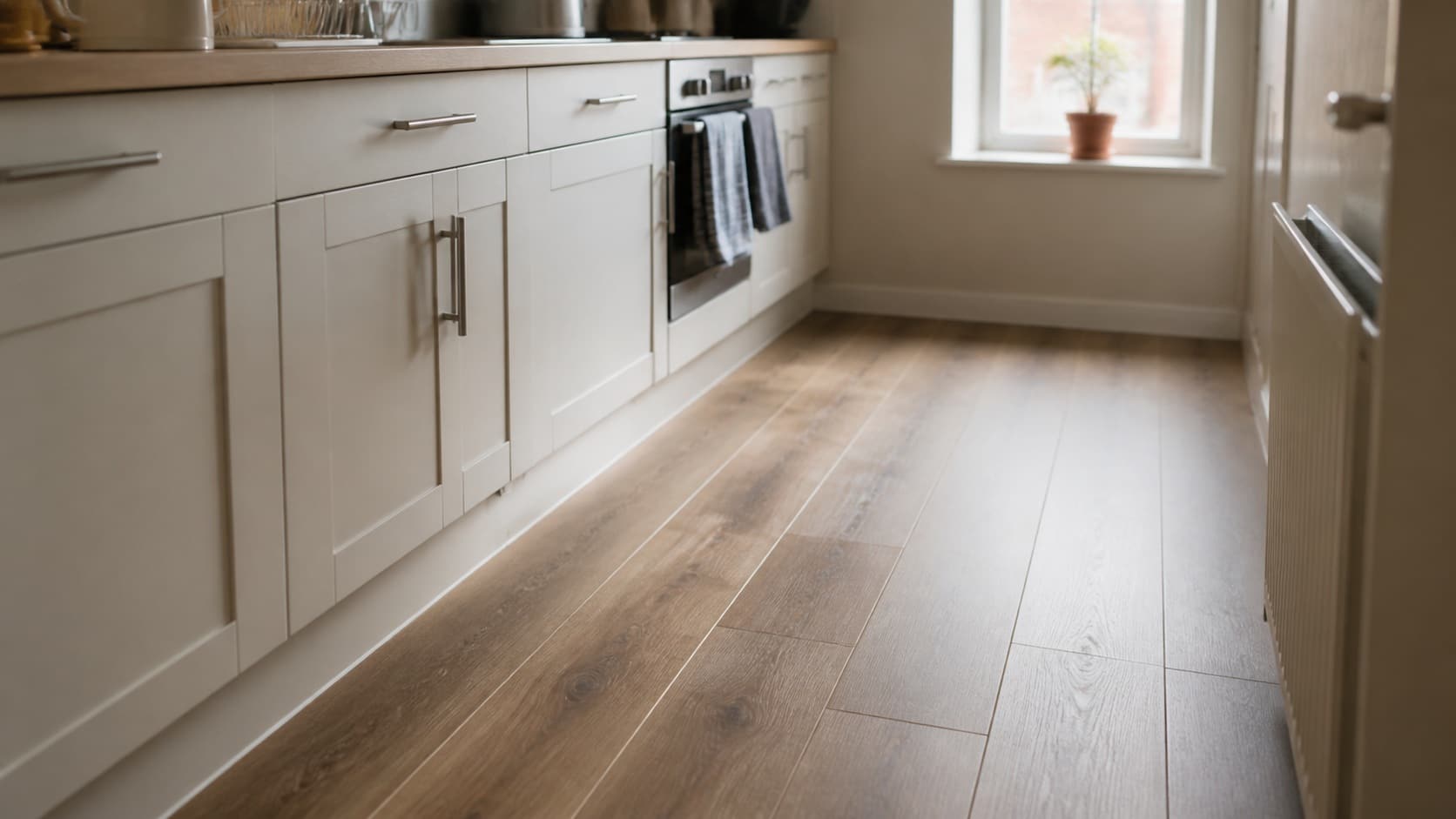 A warm oak toned luxury vinyl plank floor running under kitchen base cabinets toward a bright window
